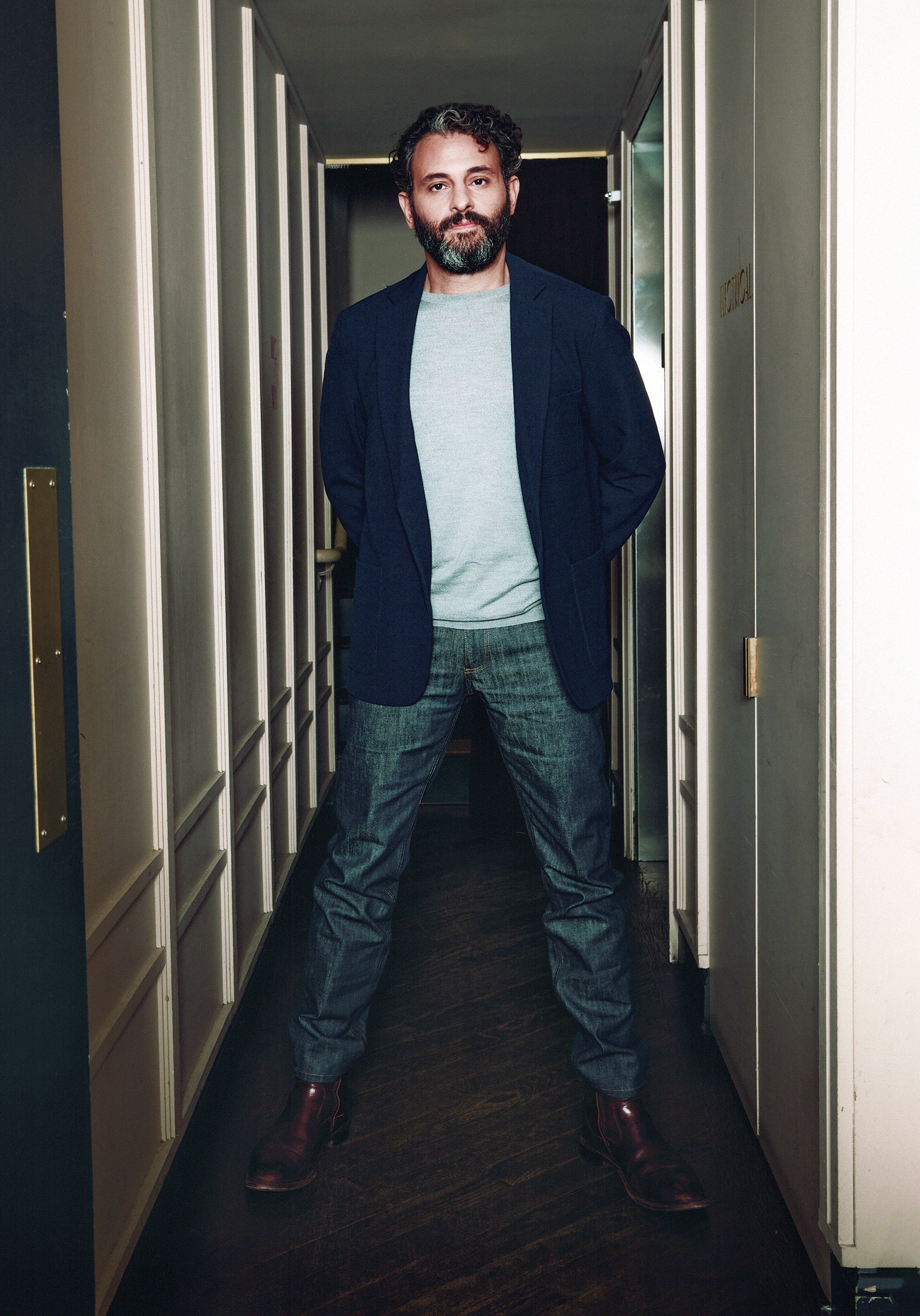 Arian Moayed wears jeans and a blue blazor posing in a long hallway