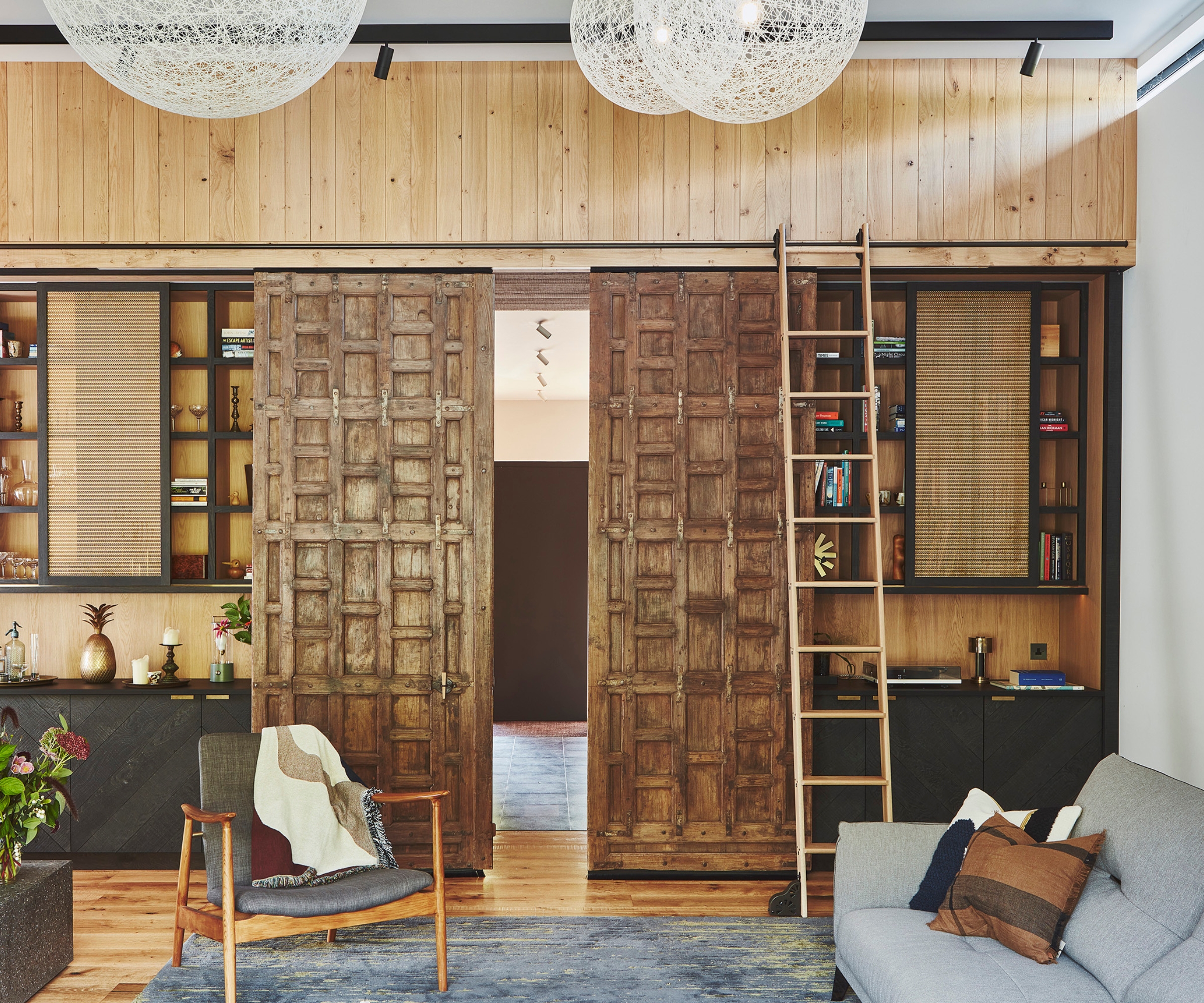 Rustic carved wooden sliding doors between shelving units, string pendant lights, grey sofa and armchair.