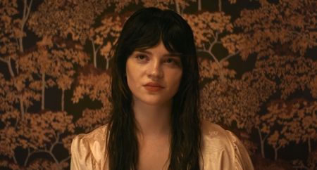 Elle sitting against backdrop of trees with yellow leaves in Her Private Hell