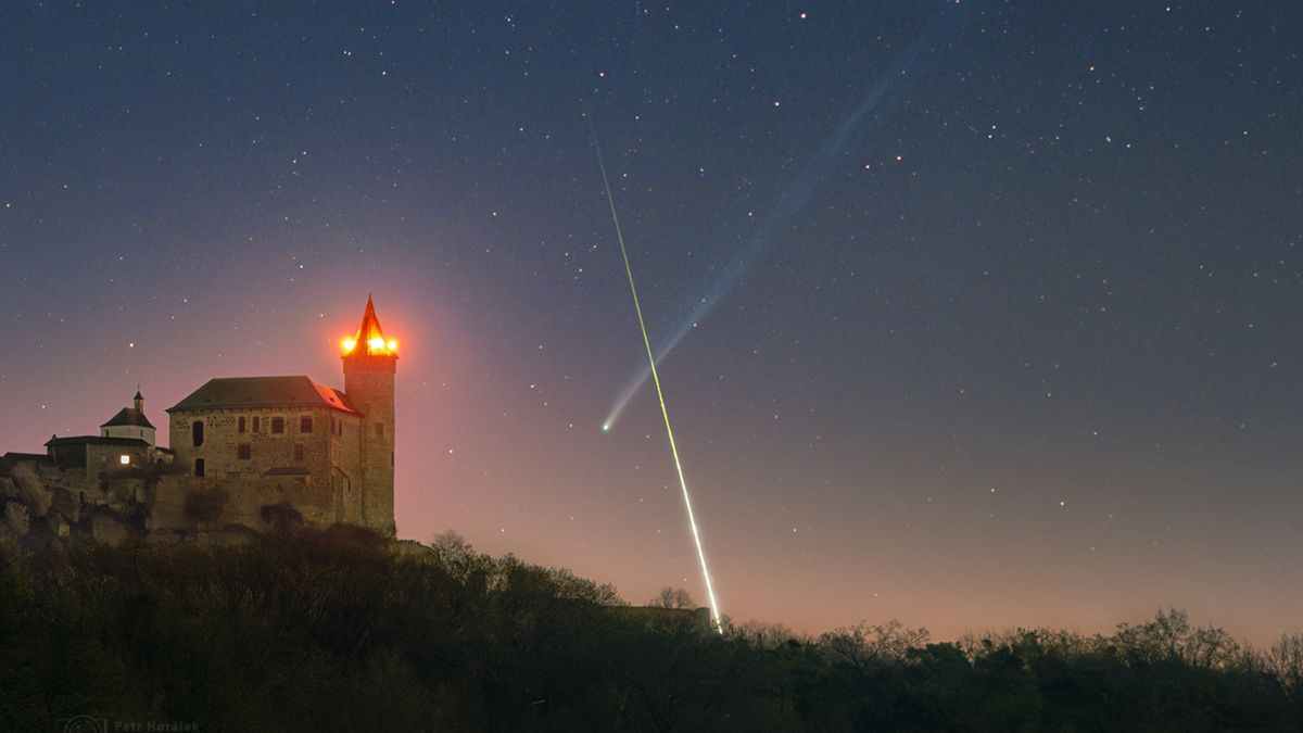 A timelapse image showing a bright streak of light crossing in front of a comet's long tail to form an X-like shape in the night sky. A medieval castle is illuminated in the foreground.