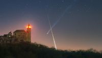 A timelapse image showing a bright streak of light crossing in front of a comet's long tail to form an X-like shape in the night sky. A medieval castle is illuminated in the foreground.