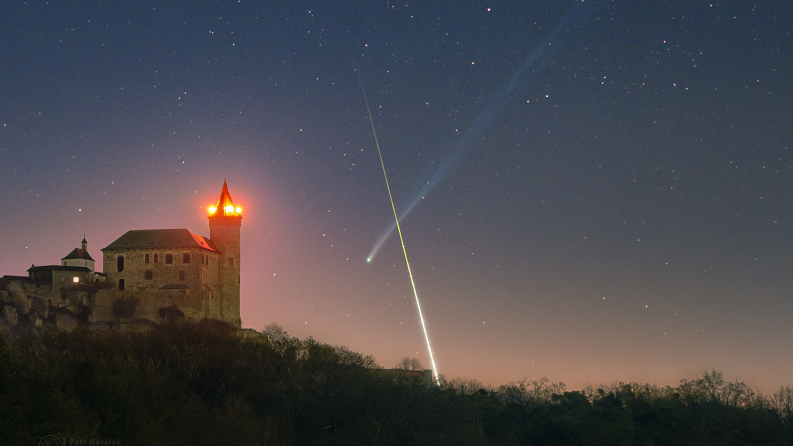 A timelapse image showing a bright streak of light crossing in front of a comet's long tail to form an X-like shape in the night sky. A medieval castle is illuminated in the foreground.