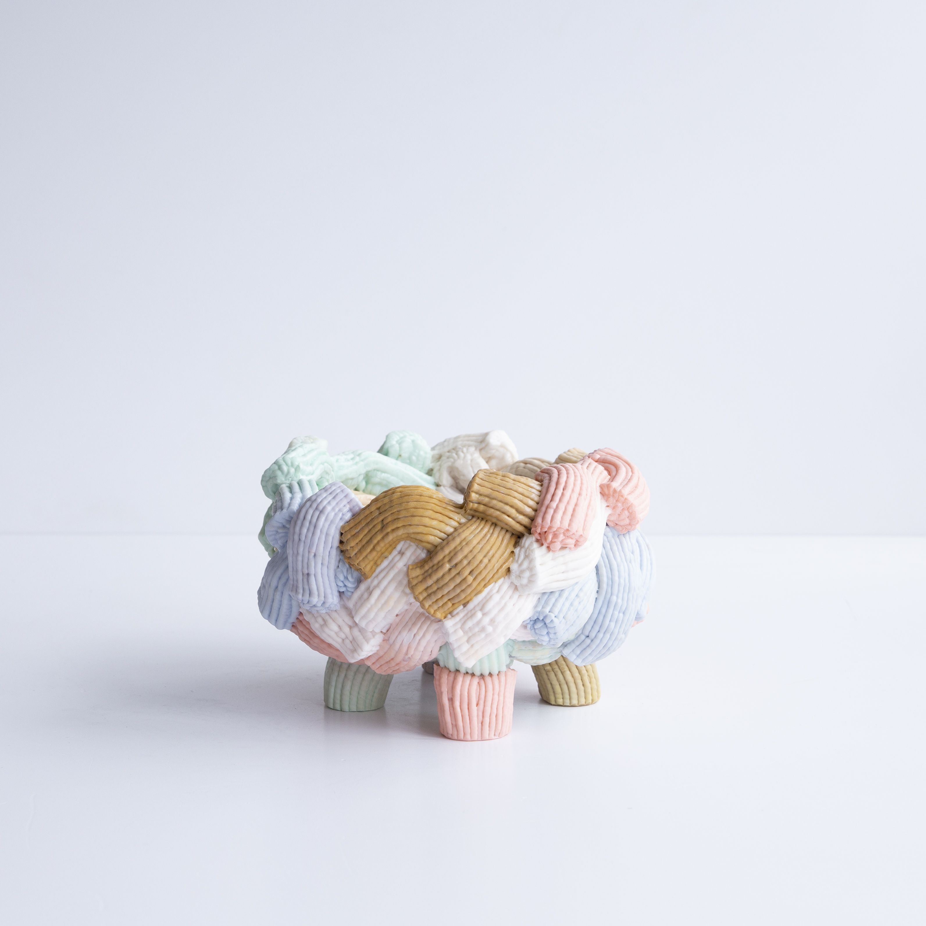 A pastel-shaded, legged vase made of multiple, embracing fragments of icing-looking plastic sits in a white room.