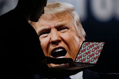 A man is seen with a laptop depicting the CNN logo with Donald Trump appearing in the background.