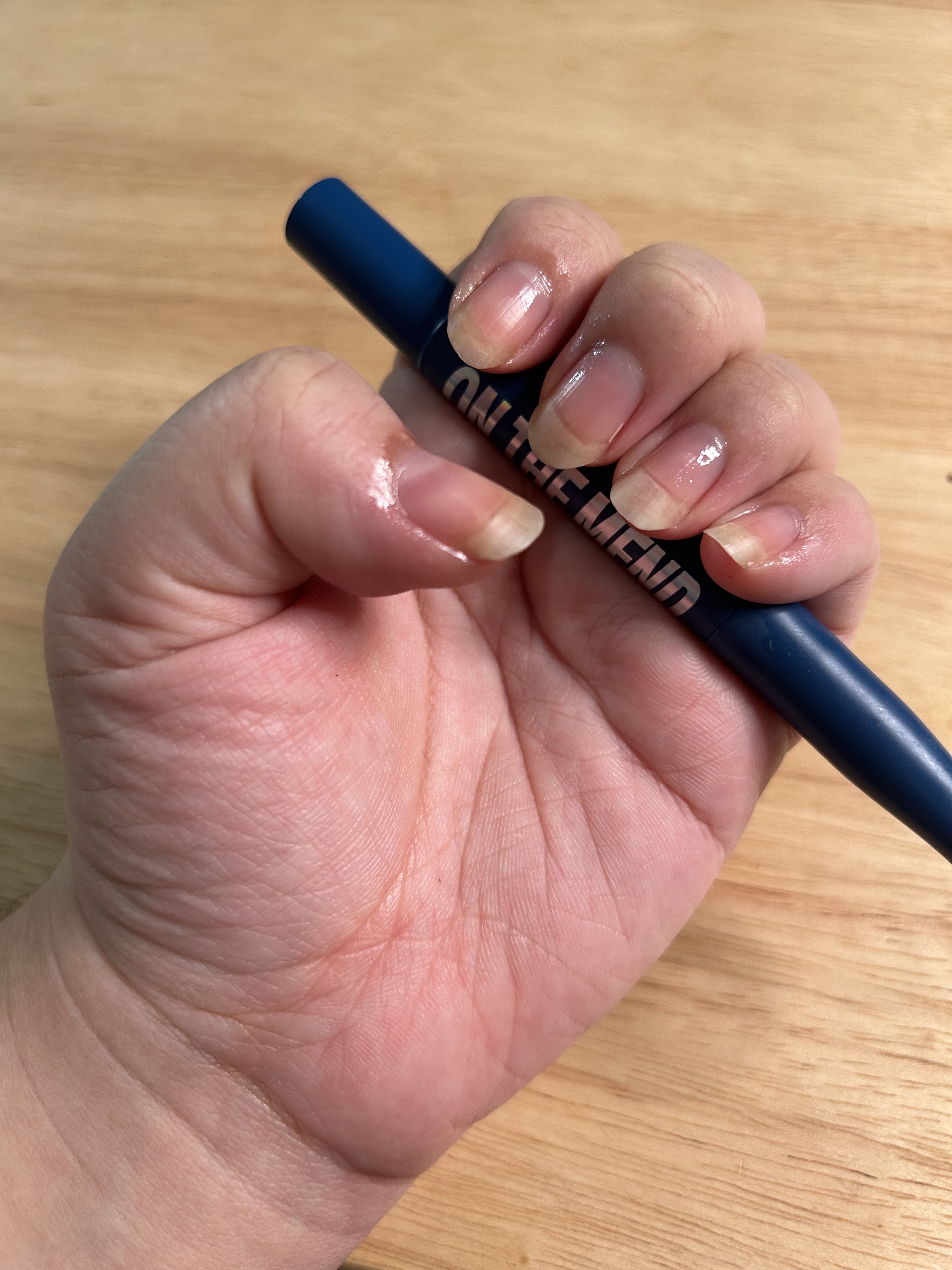 A woman's hand and nails after applying a glossy cuticle oil with a wood background and shiny nails holding the blue Chillhouse On the Mend product.