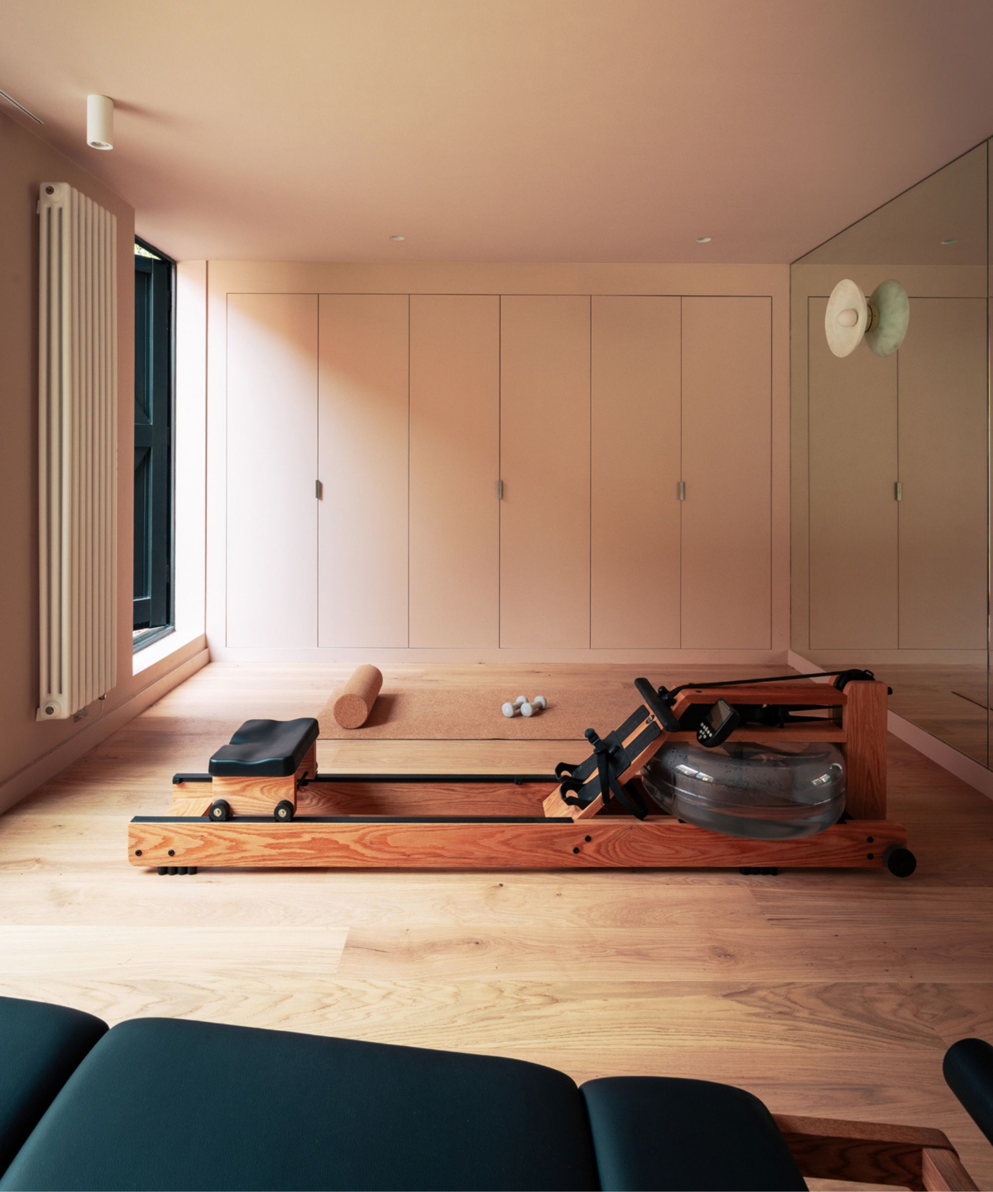 Wooden rowing machine on light hardwood floor, pink walls, built-in wardrobes, mirror, cork yoga mat, dumbbells.