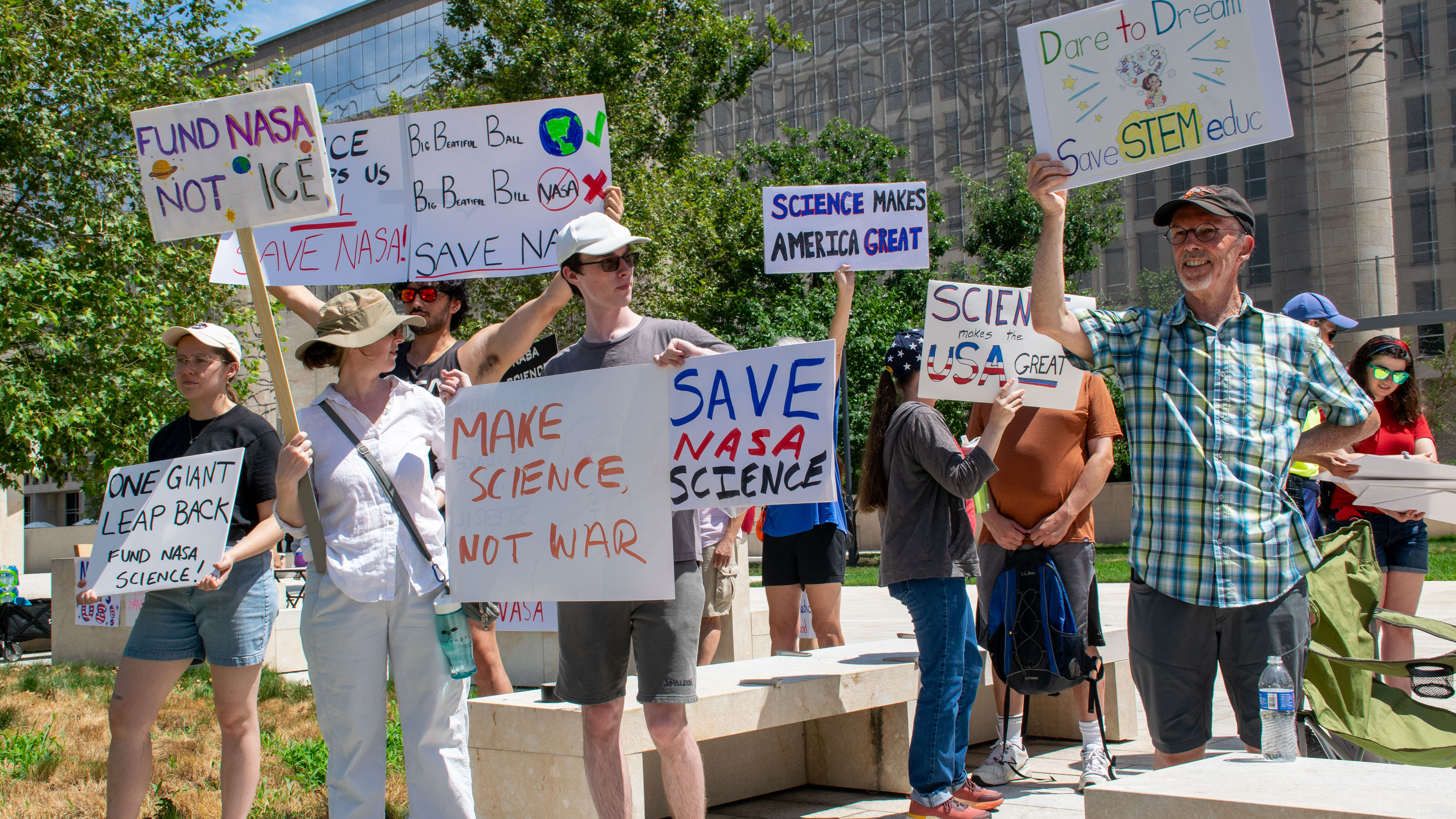 People hold signs outside on a sunny day in protest of NASA budget cuts, July 20, 2025.
