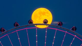 bright full moon behind a Ferris wheel with pink lights.