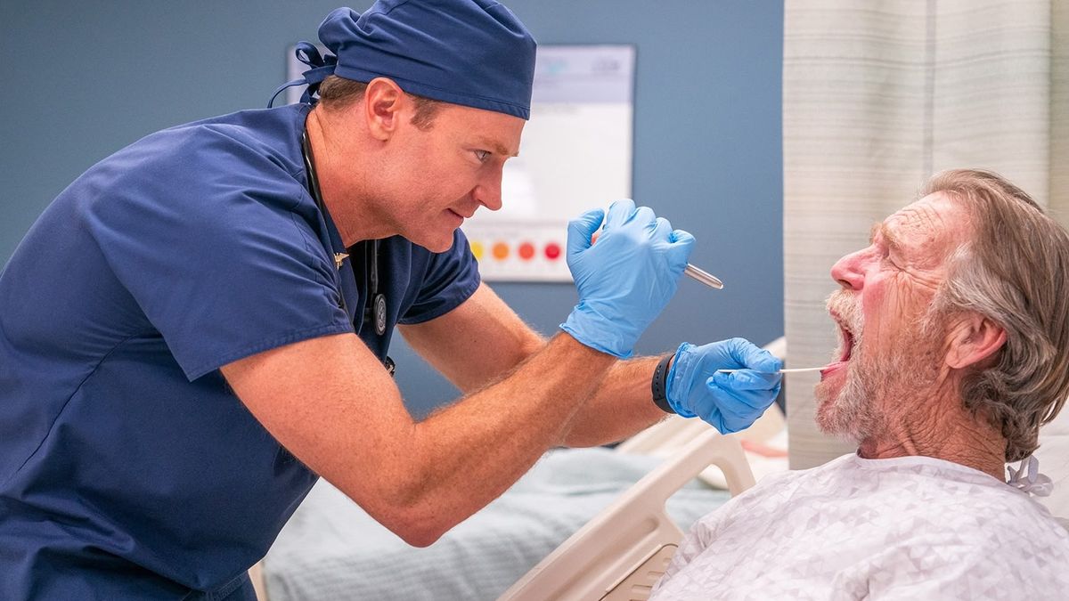 A doctor in blue scrubs comedically inspects a bed-bound patients&#039; mouth in a scene from TV show &quot;St. Denis Medical&quot; season 1