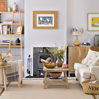 Living room with light walls and wooden pieces throughout the room ssuch as a coffee table, side table, console table, shelving and ladder leaning on the shelving, along with a woodburner fireplace in the centre of the back wall