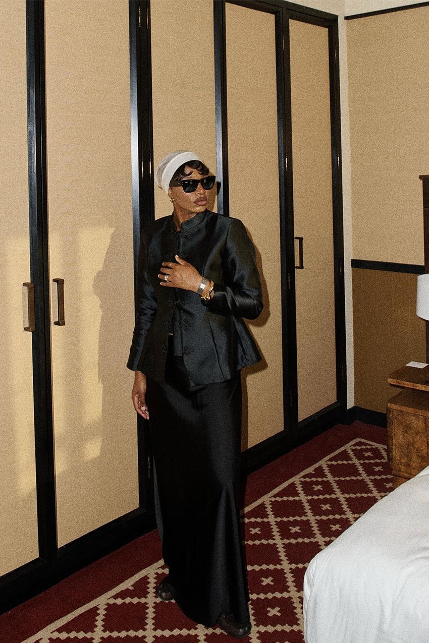 A woman standing inside a hotel room wearing a black tang jacket with matching black wide-leg trousers, black shoes, black sunglasses, gold and silver jewelry, and a white turban.