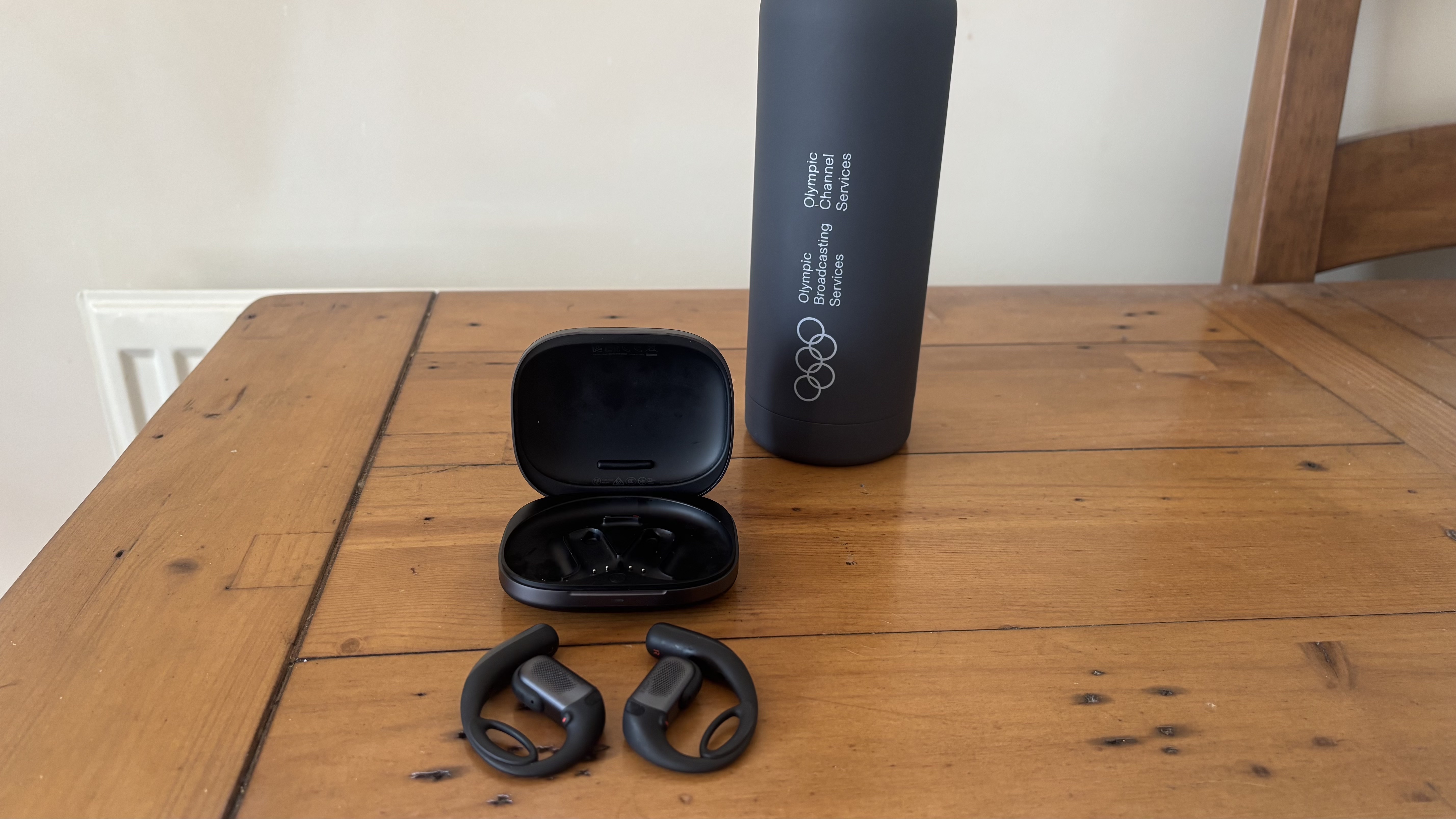 Shokz OpenFit Pro earbuds, outside their charging case, in front of a gray water bottle on a wooden surface.