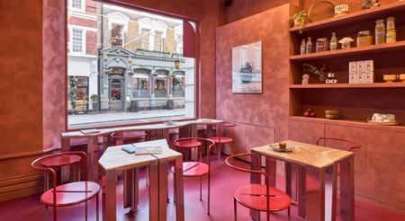 A cafe and deli with matte cement floors tinted bright pink, terracotta plaster walls, lacquered pink chairs, and marble and chrome tables overlooks an historical road.