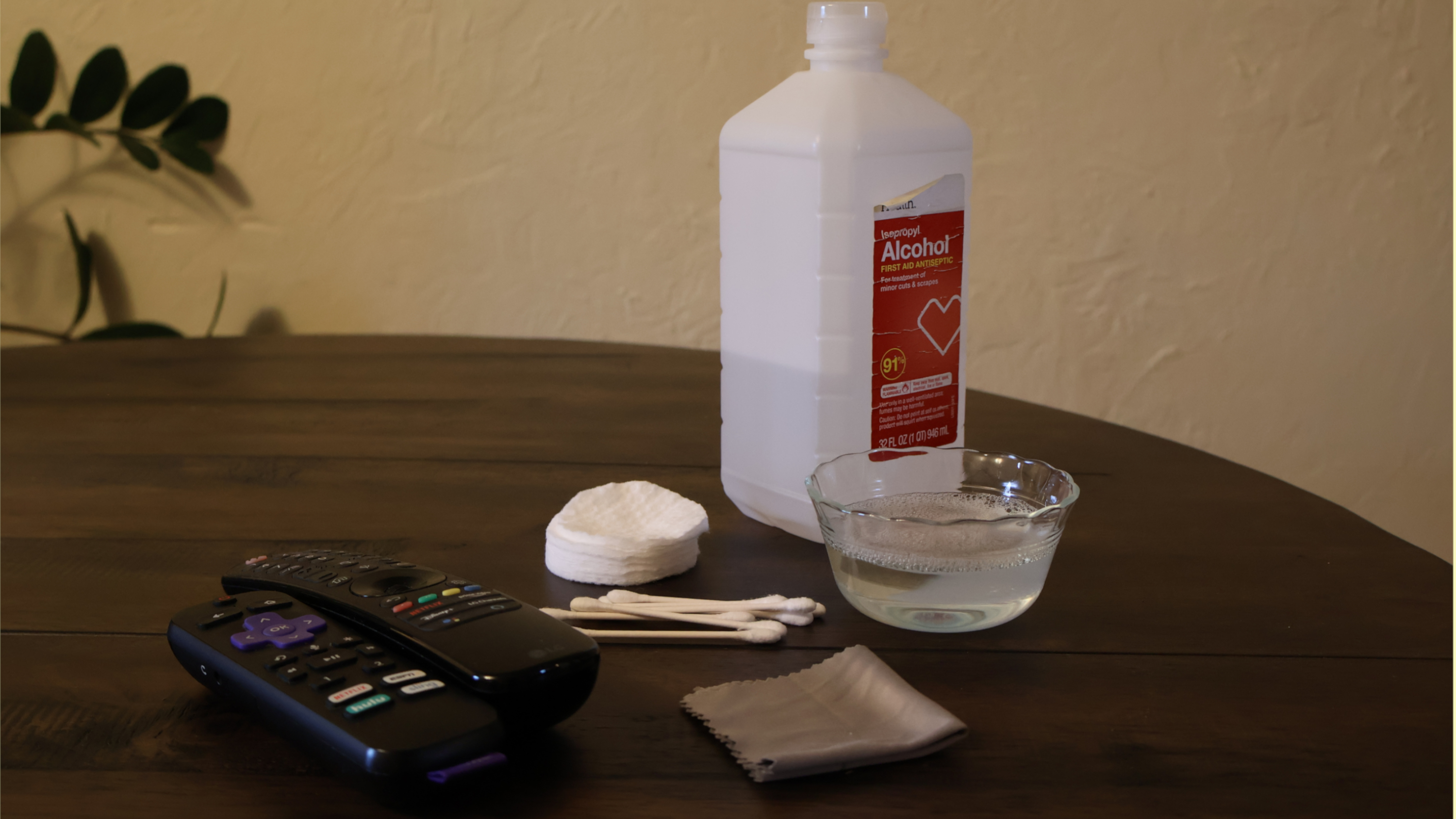 The following objects arranged on a wooden table: a bottle of isopropyl alcohol, a small stack of cotton rounds, a bundle of cotton swabs, a dish of soapy water, a folded microfiber towel and two TV remote controls