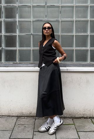 a black maxi skirt outfit showcasing a woman wearing a black waistcoat styled over a tank top with a black maxi slip skirt and white sneakers