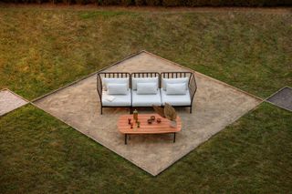 Outdoor sofa with metal body and white cushions in an outdoor setting with a long brown table dressed with a vased plant, bowls of snack, a bottle of drink and 3 glasses