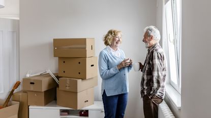 A senior couple drinks coffee with moving boxes in the background.