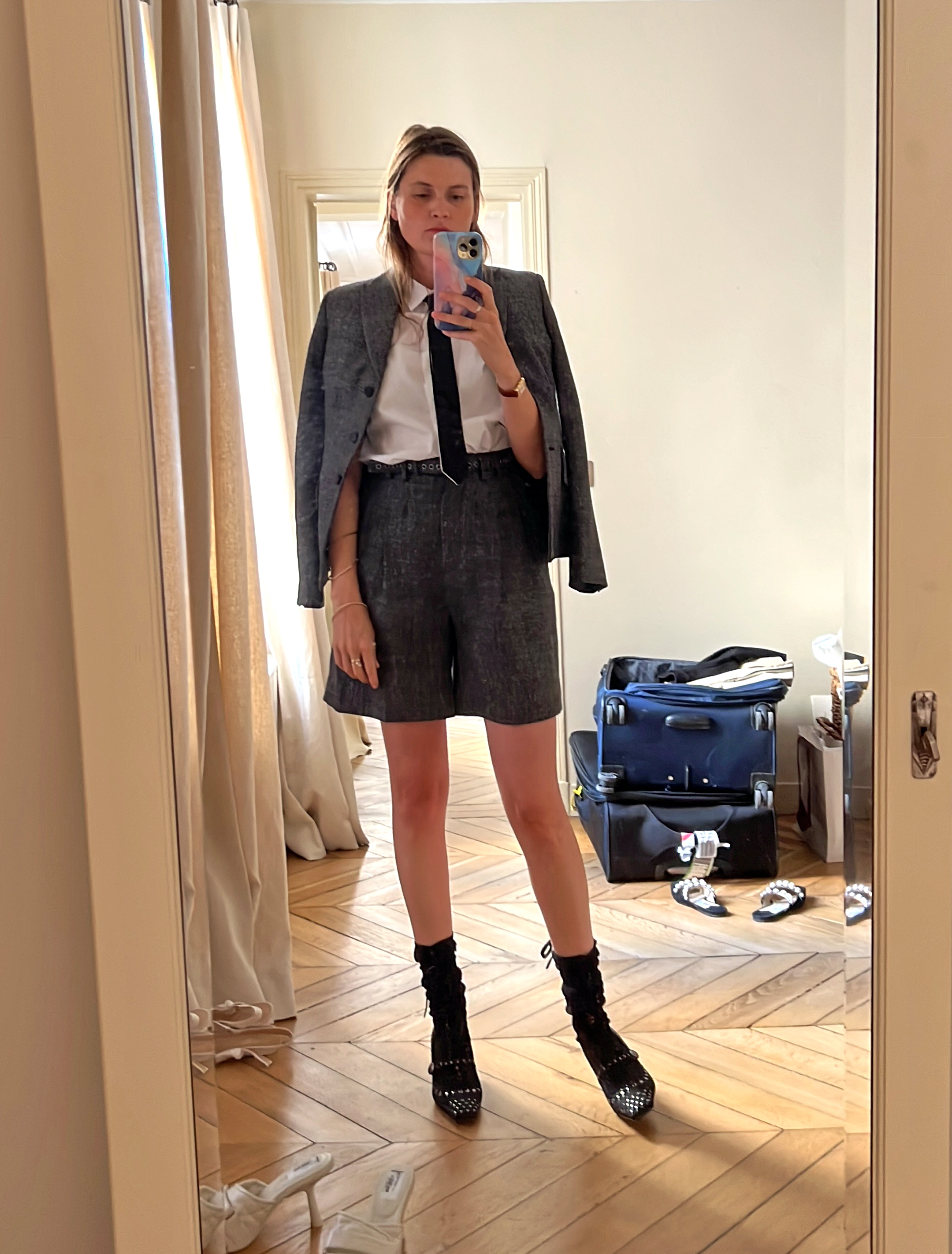 French editor and write Eugenie Trochu poses for a mirror selfie wearing a gray suit with shorts, a white button-down shirt and a tie with black socks and studded black shoes