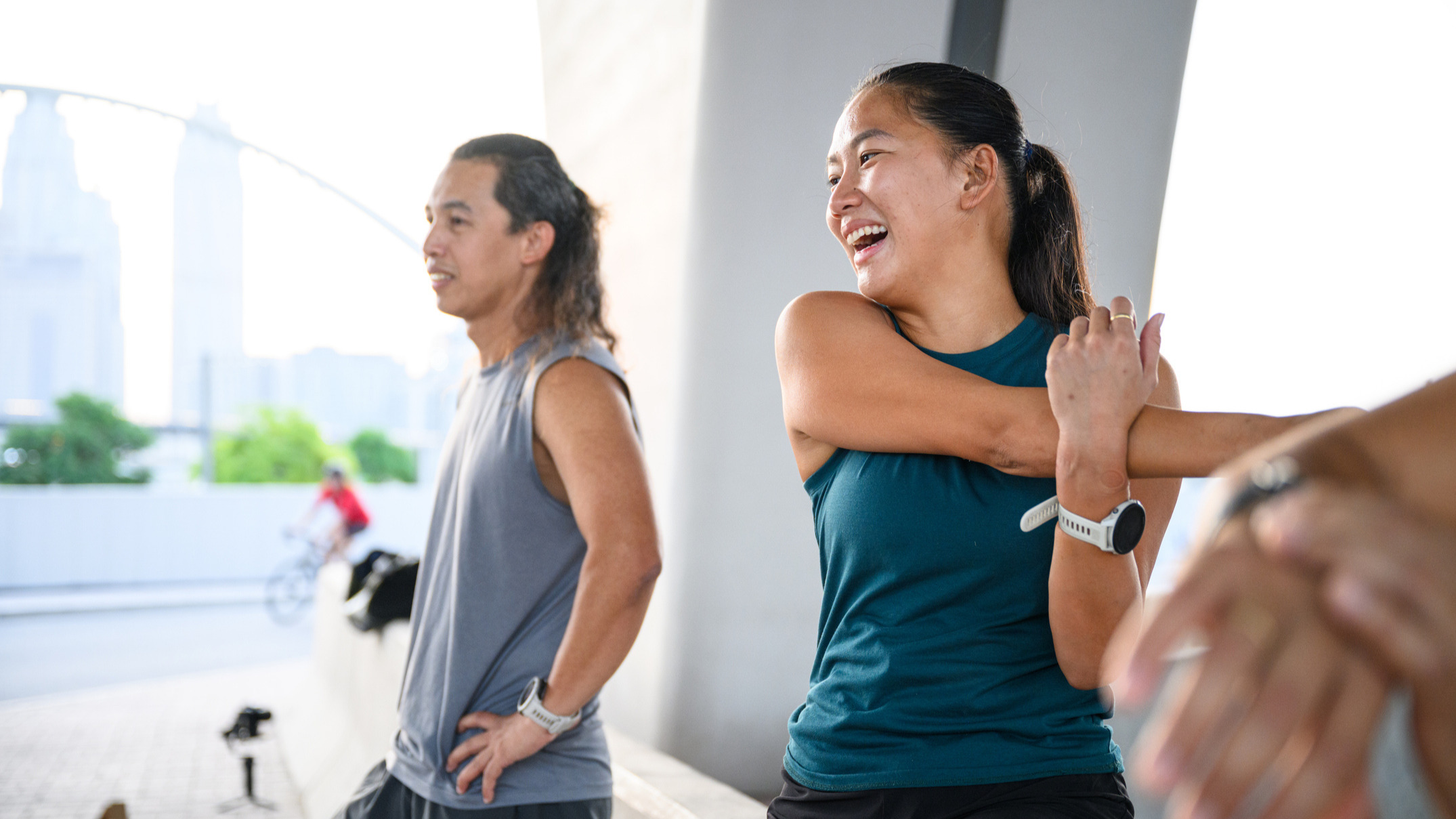A man and woman in vest tops. The woman is stretching one arm across her body and smiling with her mouth open