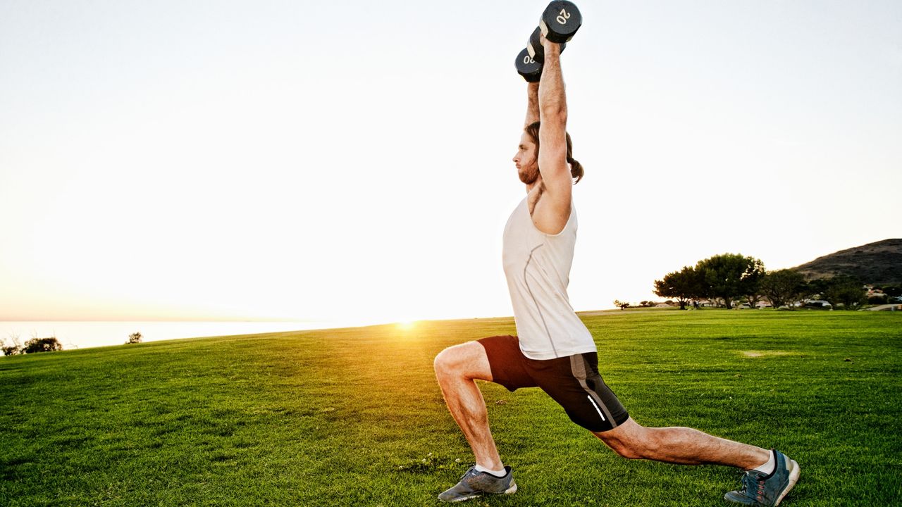 Man performing dumbbell overhead lunges on grass during outdoor workout