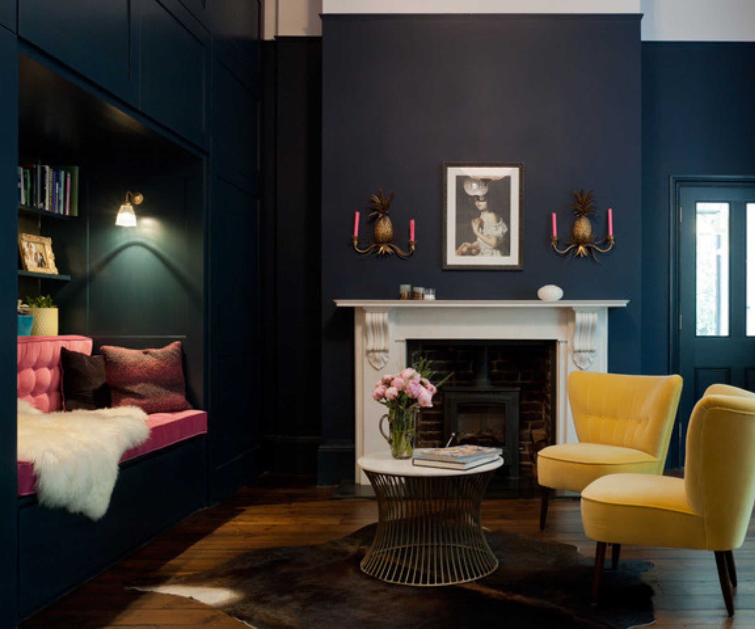 A bold, eclectic living space with deep navy walls, a white fireplace, and quirky pineapple-shaped wall sconces holding pink candles. A built-in nook with tufted pink seating and a fur throw sits opposite yellow armchairs, creating a vibrant mix of colours and textures.