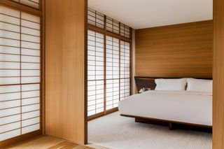 Interior view of the Tooki suite at the Shinmonzen hotel featuring wood covered walls, a bed with white pillows and linen, a sycamore headboard, tatami mat flooring and shoji screens