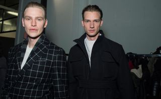 Two male models wearing looks from Canali's collection. One model is wearing a black and white jacket with grid style pattern and a white crew neck top. And the other model is wearing a black coat with pockets and a belt and a light coloured jumper