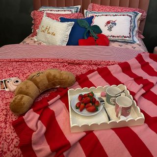 Red and pink bedsheet covers, white scalloped tray with strawberries and mugs displayed, croissant plush toy, decorative pillows