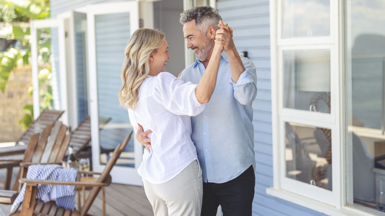 An older couple smile and laugh as they dance on the porch of their vacation home.