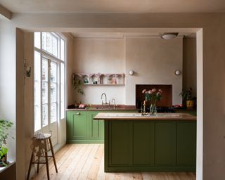 green kitchen cabinets with plaster pink walls, original floorboards, door to garden, ornate pink shelf, stool, brass faucet, wall lights, kitchen island with flowers