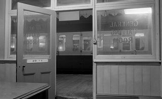 Black and white image of a train station waiting room, with the door open to the platform