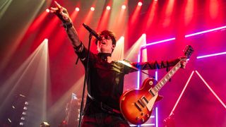Gary Numan in 2024 playing a live show dressed in black with red stage lights behind and holding a Les Paul guitar