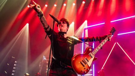Gary Numan in 2024 playing a live show dressed in black with red stage lights behind and holding a Les Paul guitar