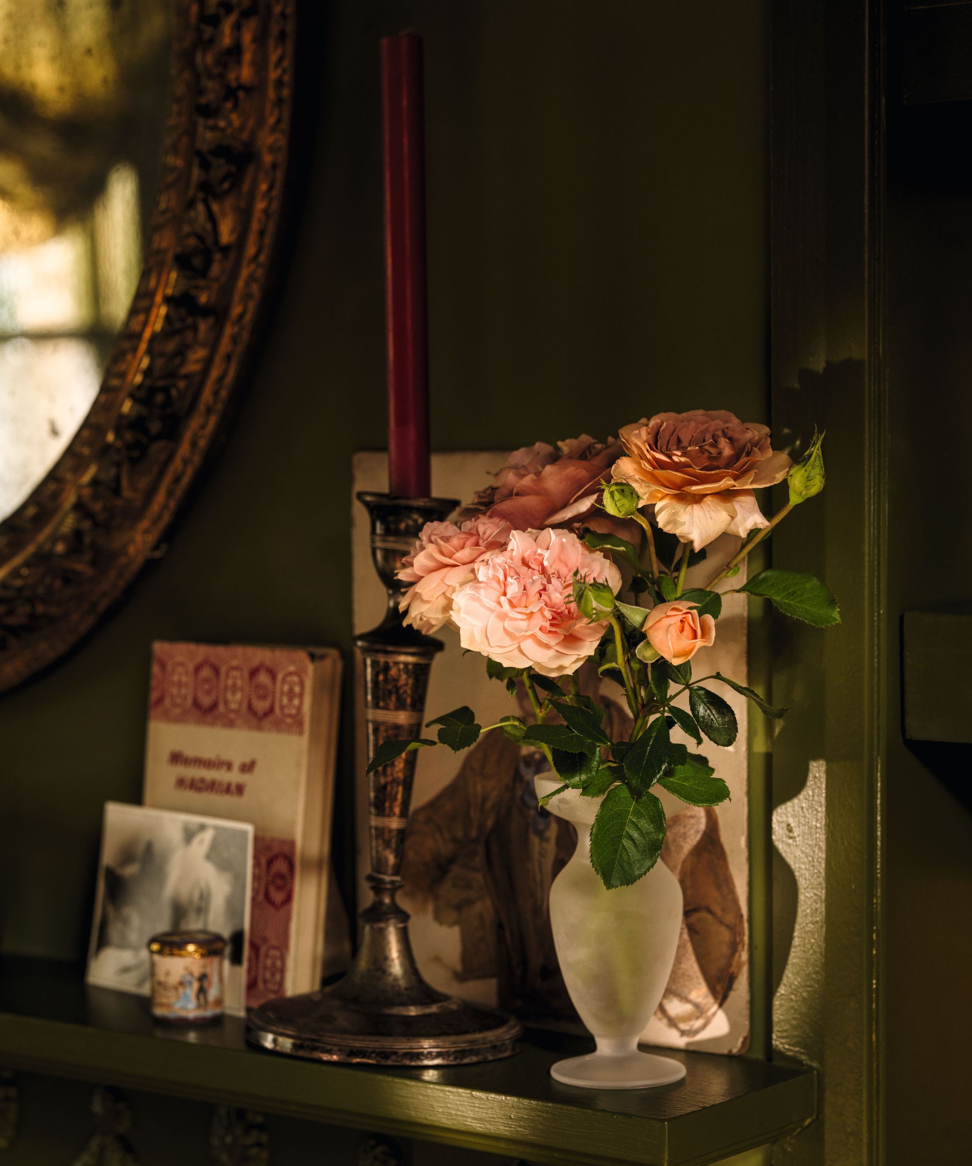 Frosted glass vase of pink roses beside an ornate silver candlestick, a red candle, vintage books, and a mirror on a dark green shelf.