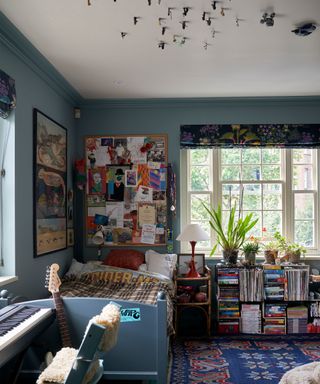 Blue boys bedroom, blue painted walls and carpet, with single bed, notice board above bed with posters and artwork, figurines stuck to ceiling, keyboard with seat, low shelving with books, patterned rug and blind