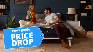 A couple sit and laugh on top of the DreamCloud Luxury Hybrid Mattress, with a blue price drop mattress sales badge overlaid on the image