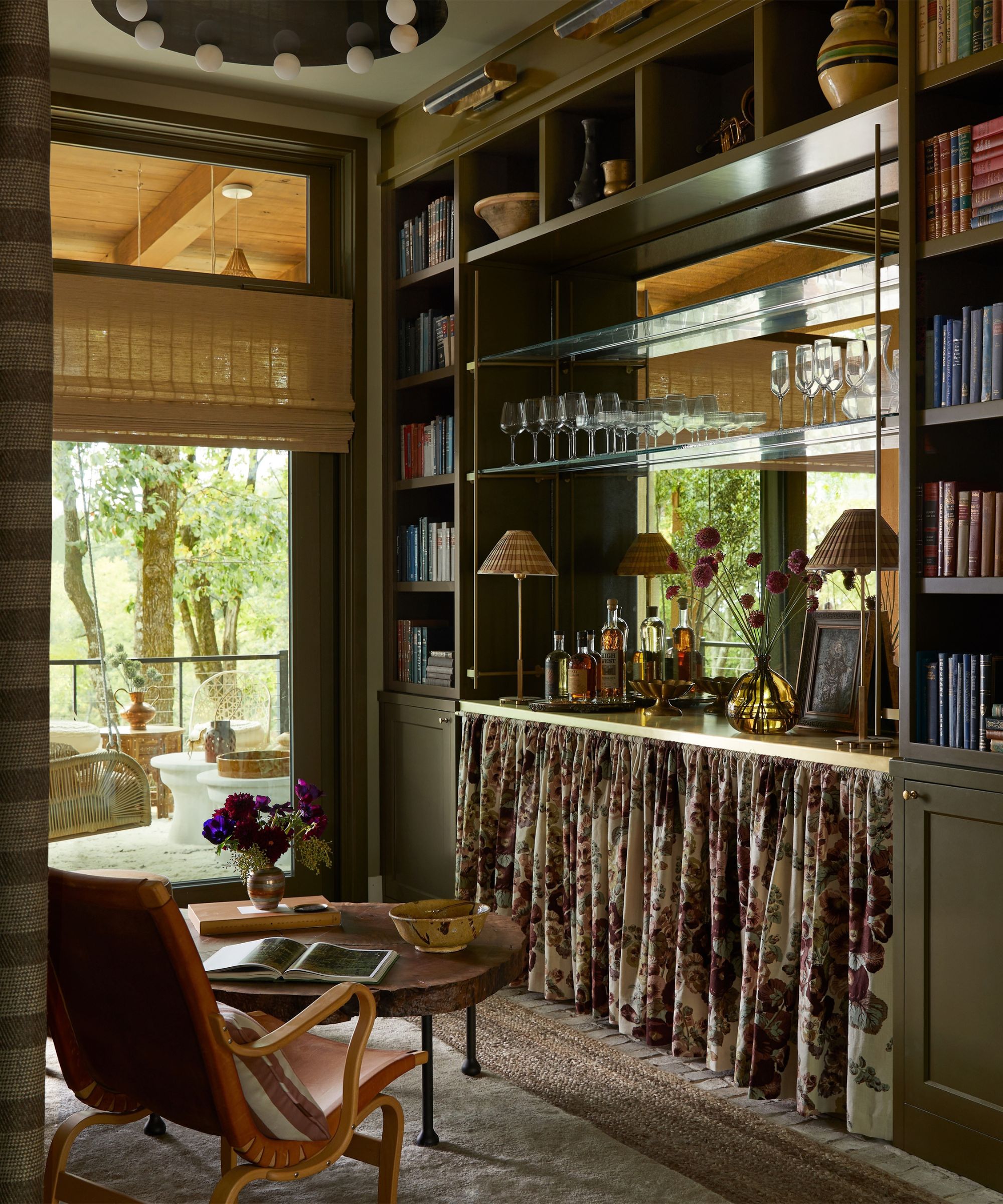 Home bar with Bancha-painted cabinetry, a floral skirted cabinet, mirrored shelving with glassware, and a wooden low table with leather armchairs, overlooking a garden with trees.