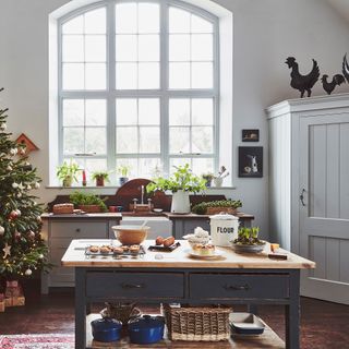 christmas baking on a farmhouse table used as kitchen island with Christmas tree and arched window and pantry cupboard behind