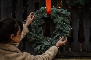 A woman hanging a pine wreath with LED string lights and a red ribbon, outside on a wooden fence.