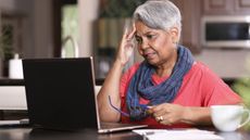 An older woman looks stressed as she looks at her laptop while at the dining room table.