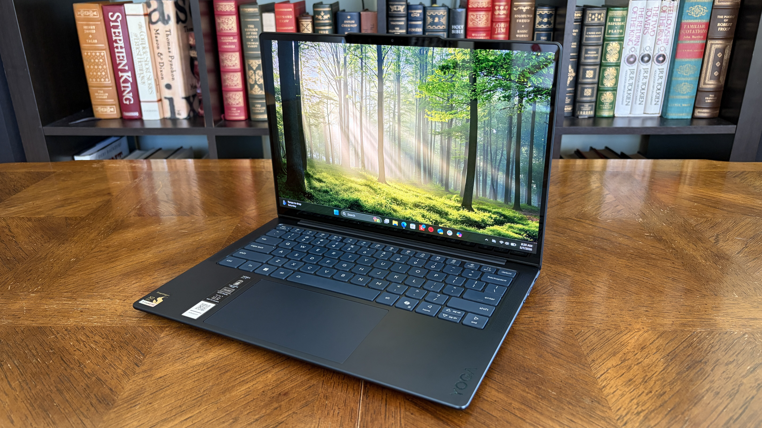 A sleek laptop with a forest wallpaper on the screen sits on a wooden table, surrounded by shelves filled with colorful books, creating a serene, studious ambiance.