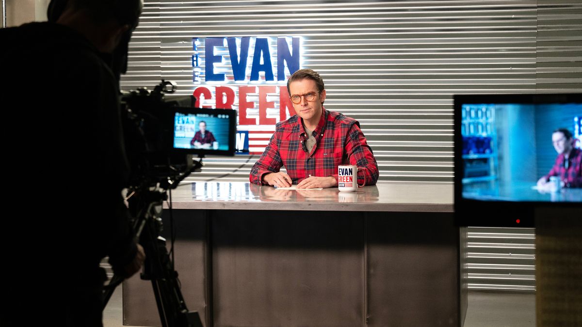 Dan Stevens as Evan Green dressed in a red and black shirt sitting at a desk against a metallic background with a sign in blue and red of his name, being filmed for his TV show. 