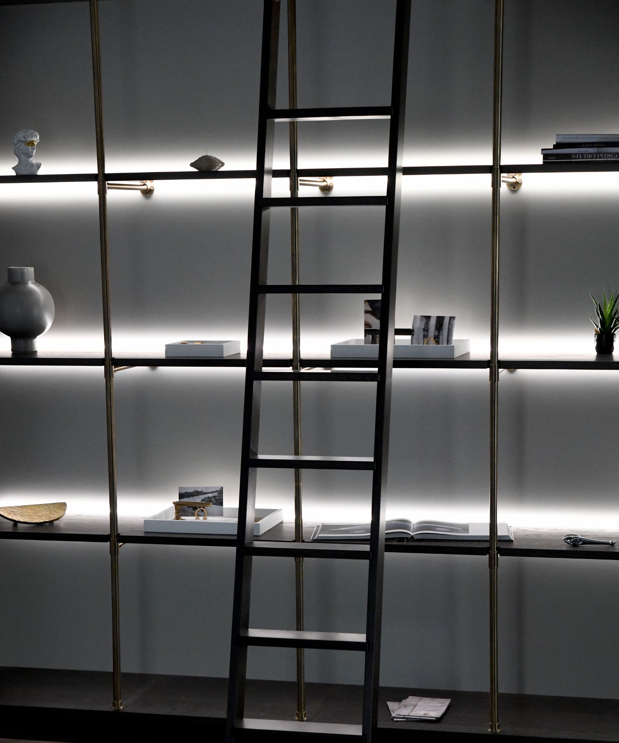 A tall, modern shelving unit features thin brass supports and dark horizontal shelves, evenly spaced across the height of the wall. Integrated LED backlighting runs the width of each shelf, casting a soft, white glow against the grey wall behind.