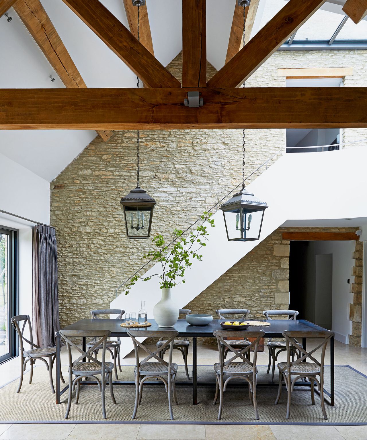 Dining area with exposed stone wall, vaulted wood-beamed ceiling, steel table, gray wood chairs, hanging lanterns