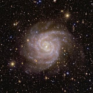A spiral galaxy is seen in the center of the image. In the background, a wealth stars and galaxies in space.