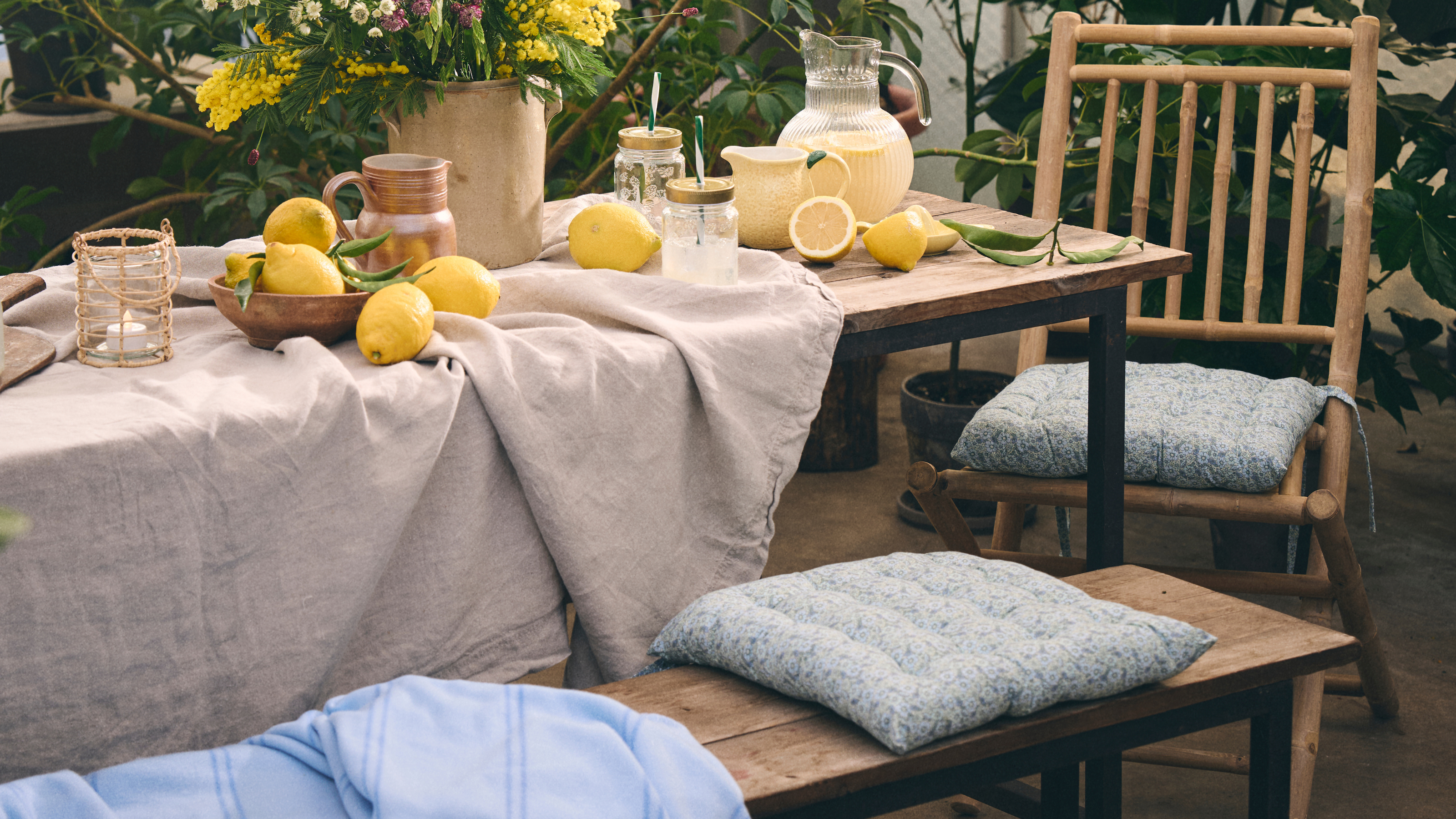 Wooden bench and chairs with neutral tablecloth, jugs of lemondade, bowls of lemons and a large bunch of flowers.