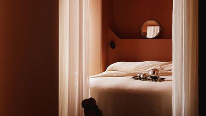 A moody bedroom with dark terracotta walls and creamy white bedding. There are white sheer curtains hanging in front of the bed, and a silver tray on the bed.