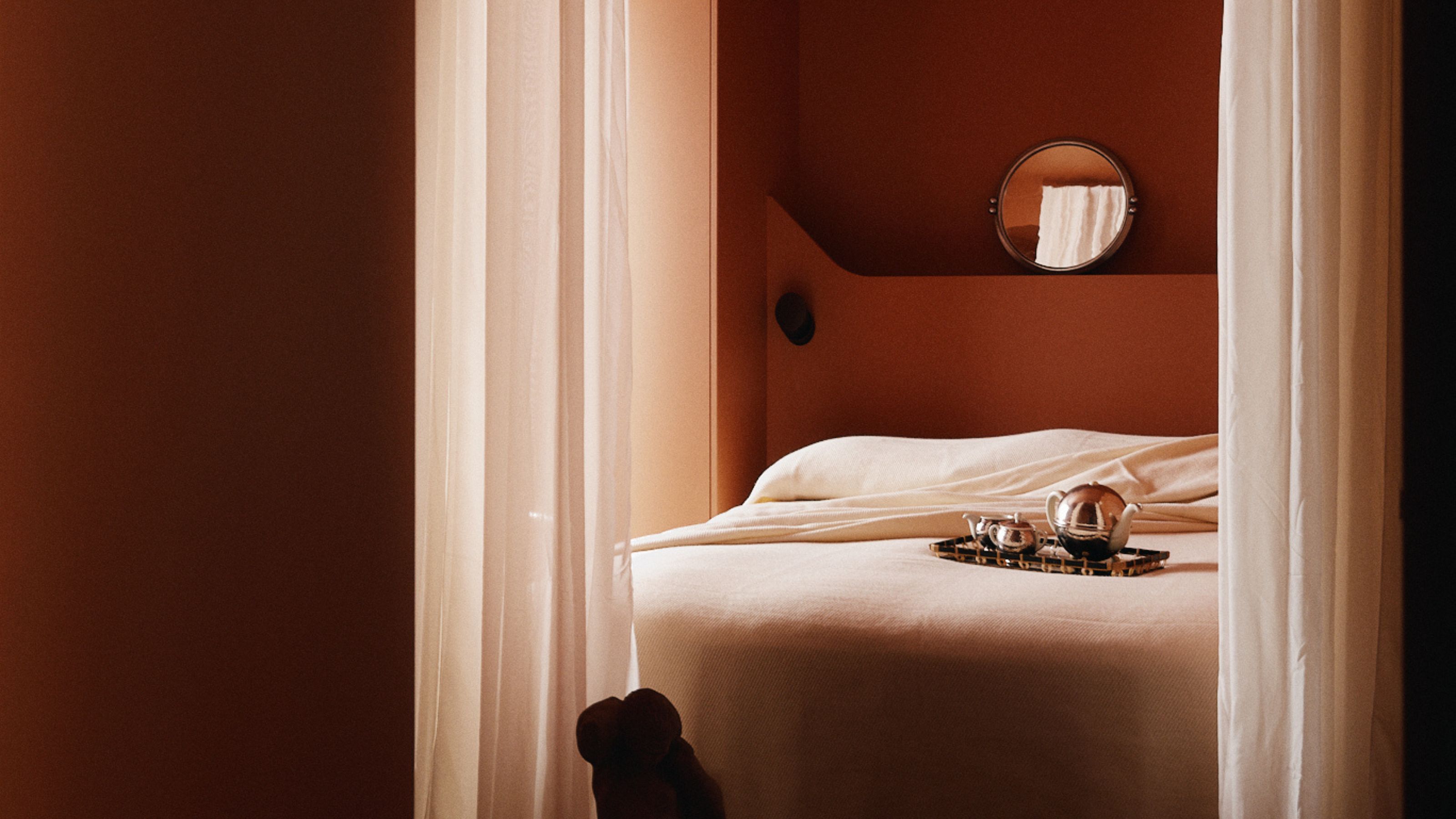 A moody bedroom with dark terracotta walls and creamy white bedding. There are white sheer curtains hanging in front of the bed, and a silver tray on the bed.