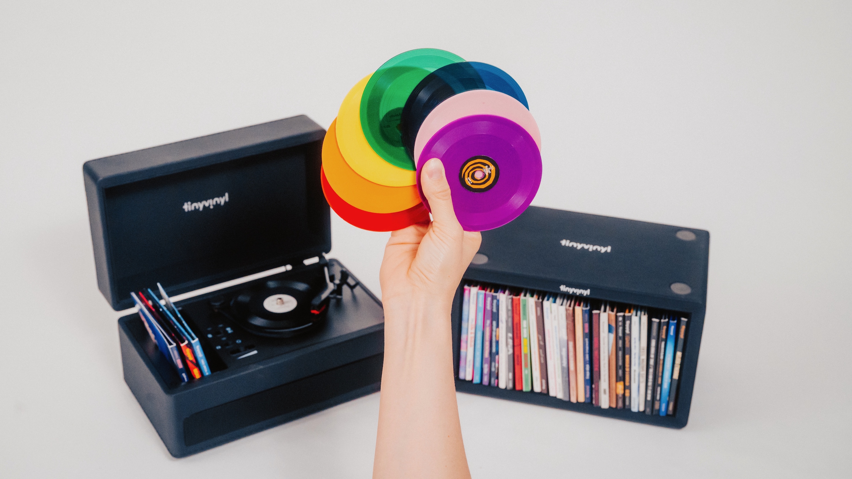 A hand fanning out Tiny Vinyl 4-inch records between the Tiny Vinyl Player turntable and Crate storage unit.
