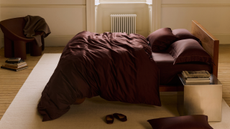 A deep burgundy cotton sateen duvet set from Rise & Fall styled on a bed in the middle of a white bedroom beside a steel side table